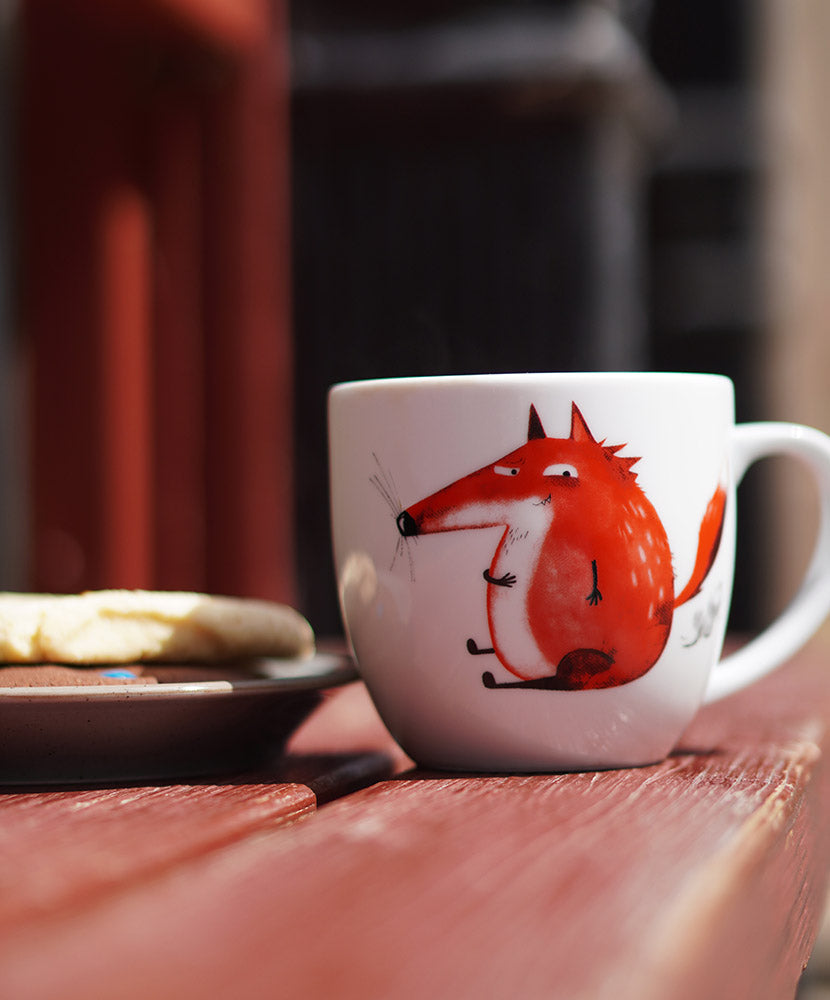 Tasse mit einem dicken pupsenden Fuchs drauf steht auf einer roten Bank.